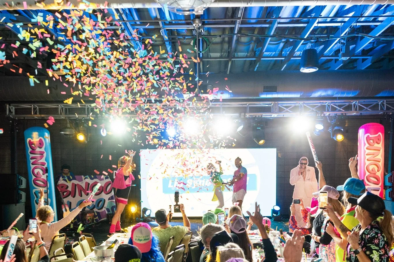Blue light washing the ceiling and stage lights shining over a crowded event room as confetti falls